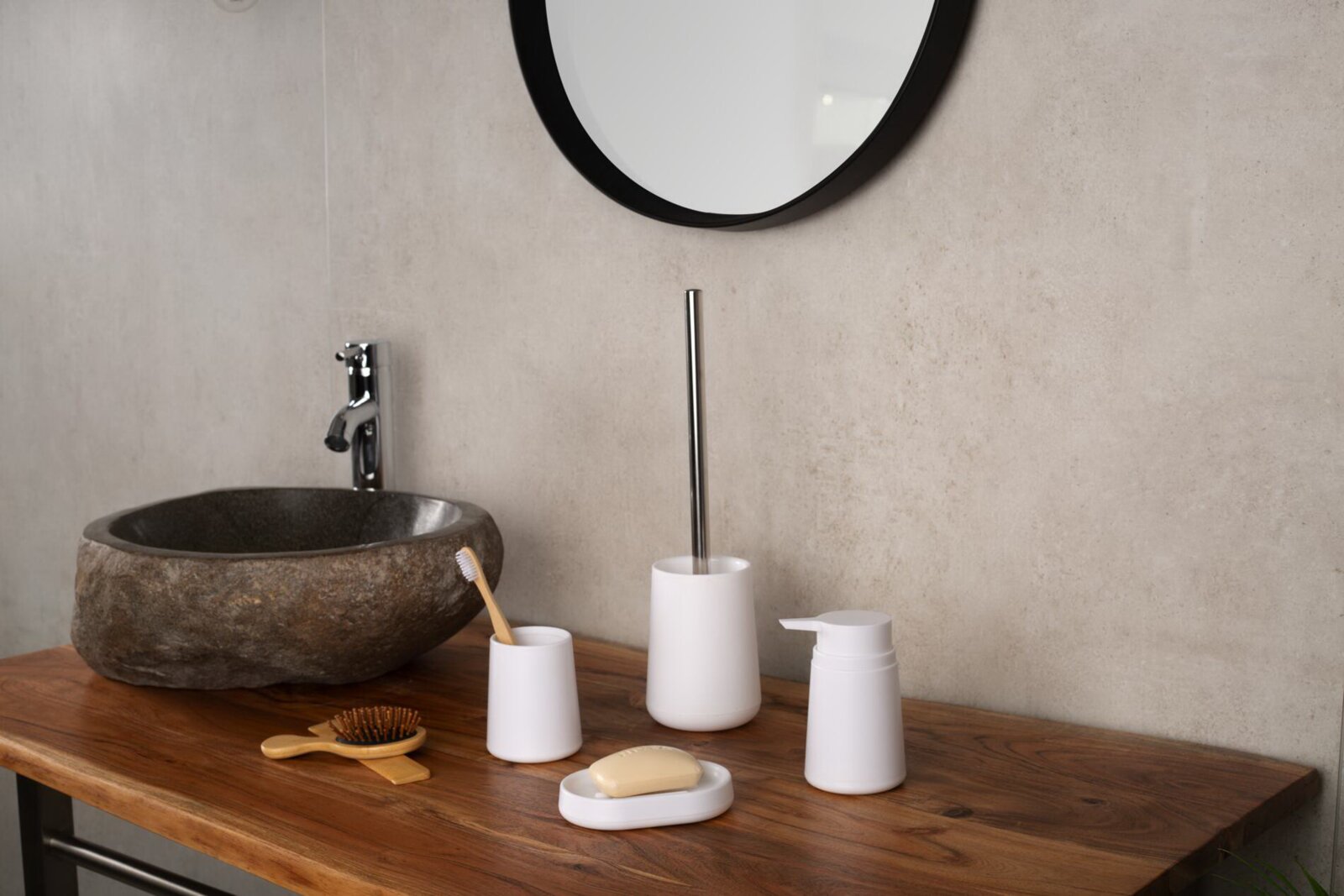 Minimalistisches Bad-Accessoire-Set in Weiß (Seifenspender, Seifenschale, Zahnputzbecher und WC-Bürste) auf Holz-Waschtisch neben Steinwaschbecken, frontale Perspektive auf Augenhöhe.
