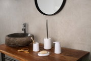 Minimalistisches Bad-Accessoire-Set in Weiß (Seifenspender, Seifenschale, Zahnputzbecher und WC-Bürste) auf Holz-Waschtisch neben Steinwaschbecken, frontale Perspektive auf Augenhöhe.