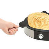 Nahaufnahme einer Hand, die einen Crêpe mit einem Spatel von der WMF LONO Creperie abhebt. Die Creperie ist aus Edelstahl mit einem Temperaturregler an der Vorderseite.