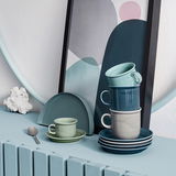 Stapel von Tassen in verschiedenen Grautönen und Blau, auf einem Tisch mit dekorativem Hintergrund, seitliche Perspektive