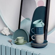 Stapel von Tassen in verschiedenen Grautönen und Blau, auf einem Tisch mit dekorativem Hintergrund, seitliche Perspektive