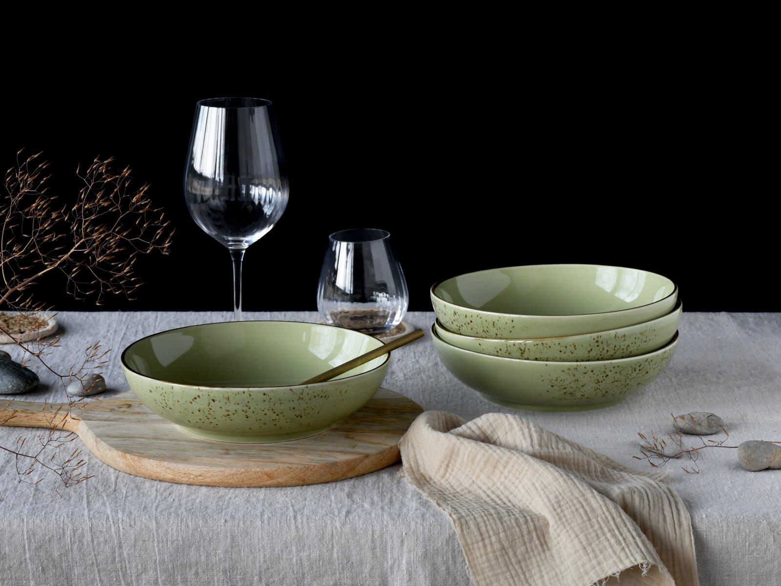 Drei gesprenkelte, salbeigrüne Keramikschalen auf einem gedeckten Tisch mit Holzbrett und Glas, fotografiert aus leicht erhöhter Frontperspektive.