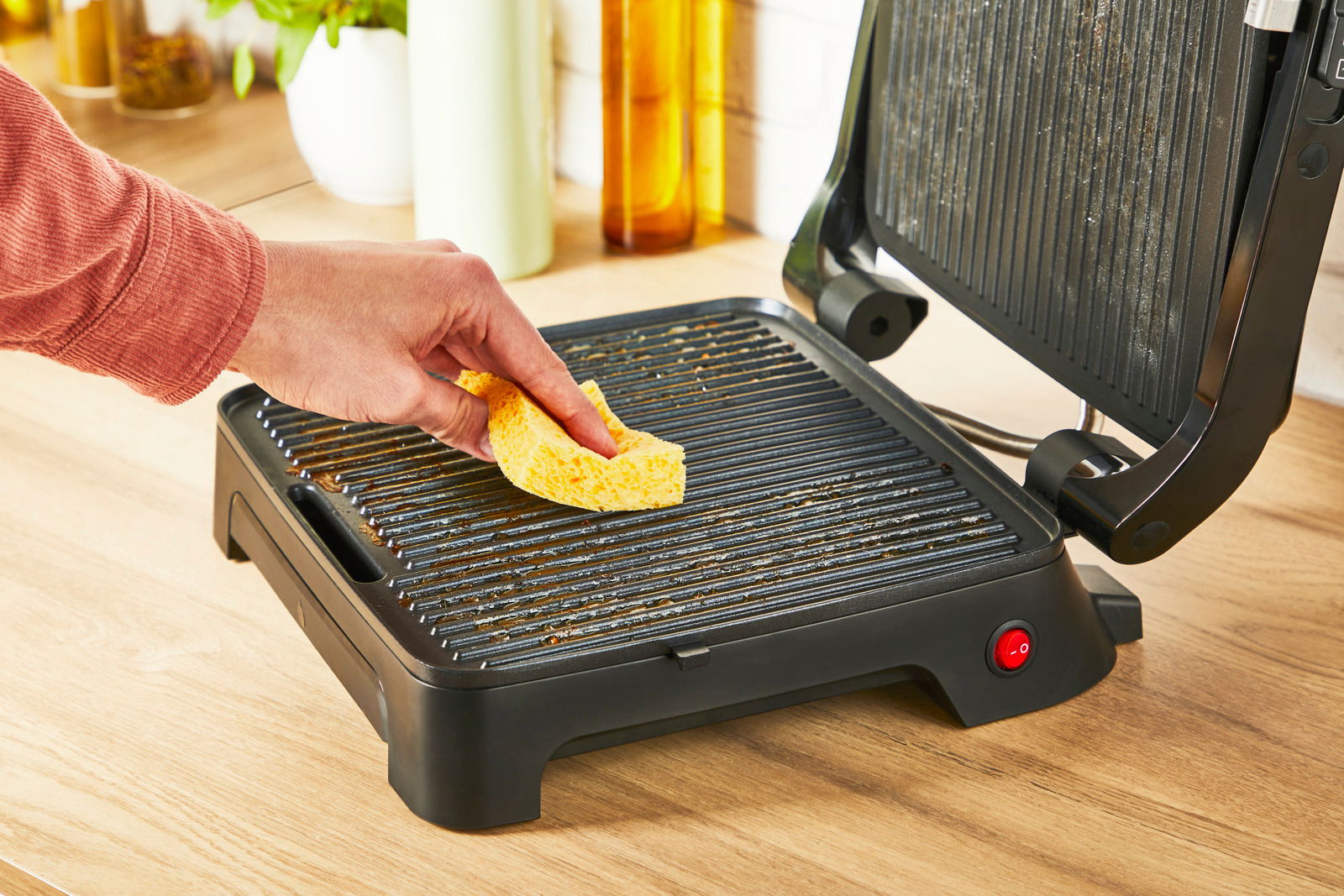 Nahaufnahme eines geöffneten Kontaktgrills Classic GC271D auf einer Küchenarbeitsplatte, während eine Hand mit einem gelben Schwamm die Grillplatte reinigt.