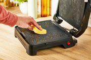 Nahaufnahme eines geöffneten Kontaktgrills Classic GC271D auf einer Küchenarbeitsplatte, während eine Hand mit einem gelben Schwamm die Grillplatte reinigt.