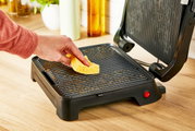 Close-up van een geopende Classic GC271D-contactgrill op een keukenblad, terwijl een hand de grillplaat schoonmaakt met een gele spons.