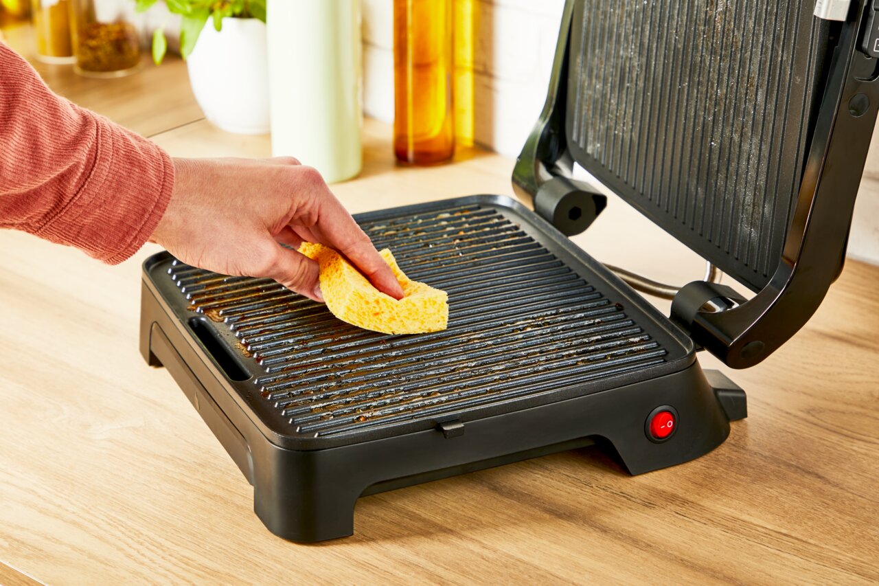 Close-up van een geopende Classic GC271D-contactgrill op een keukenblad, terwijl een hand de grillplaat schoonmaakt met een gele spons.