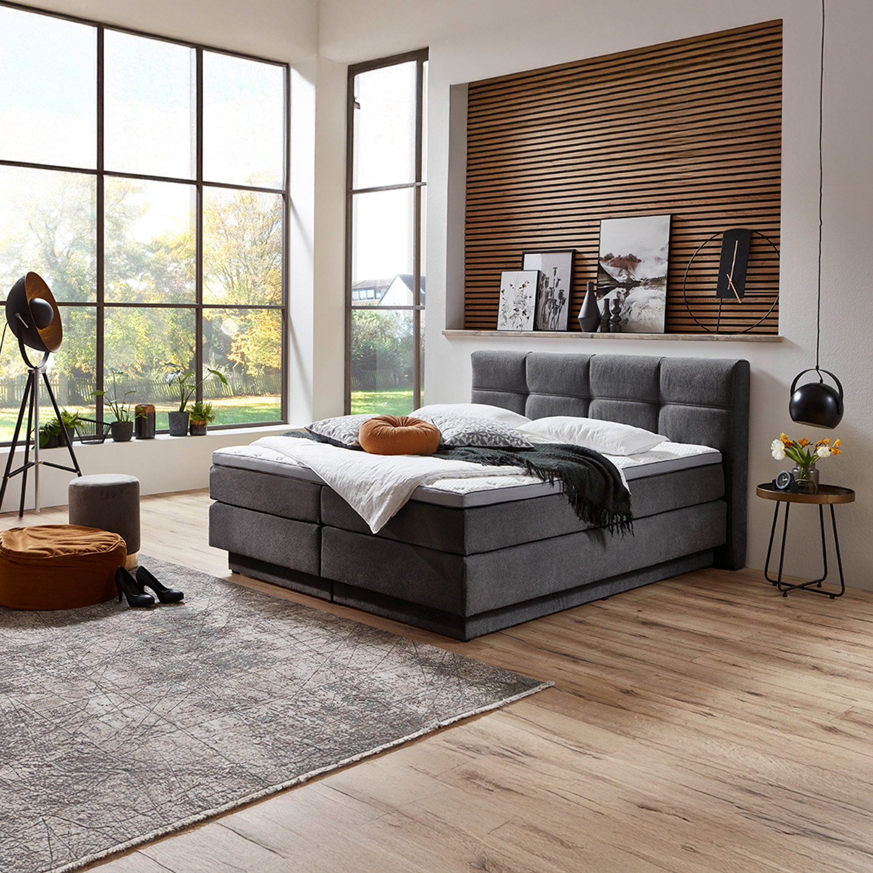 Modern eingerichtetes Schlafzimmer mit grauem Boxspringbett, dekorativer Holzlamellen-Wand, großen Fensterfronten und Blick in den Garten.