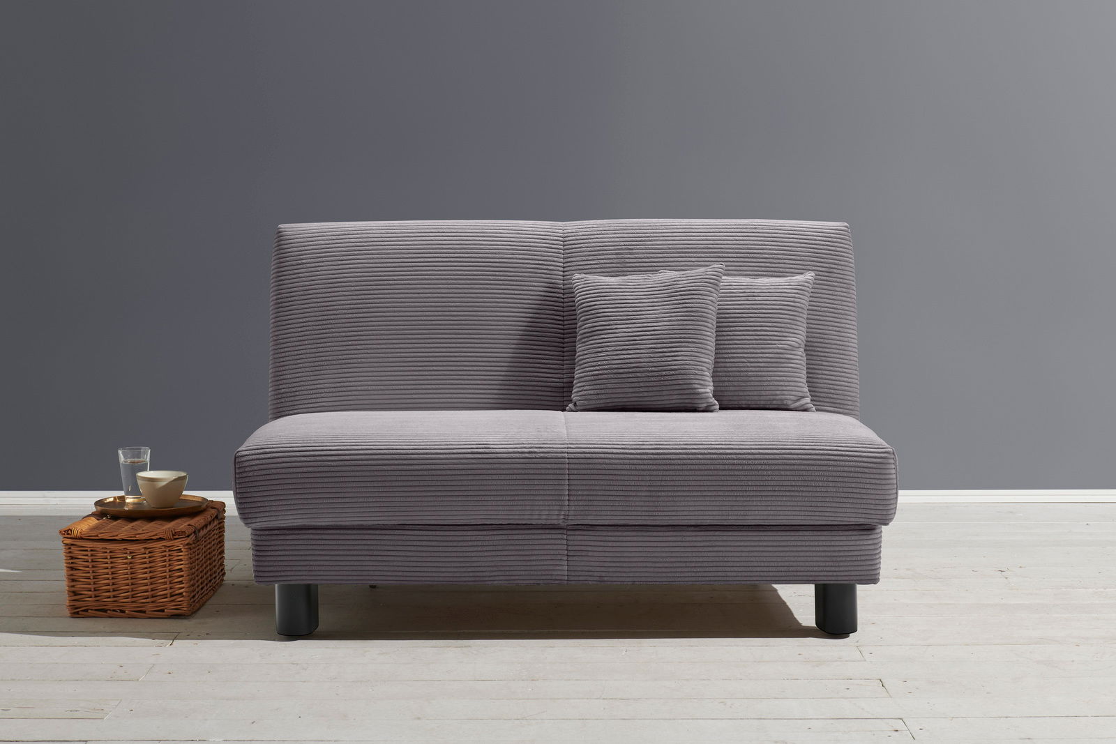 Z2 Schlafsofa ENNY Graues Funktionssofa mit zwei Kissen, frontal fotografiert, auf hellem Holzboden vor grauer Wand.