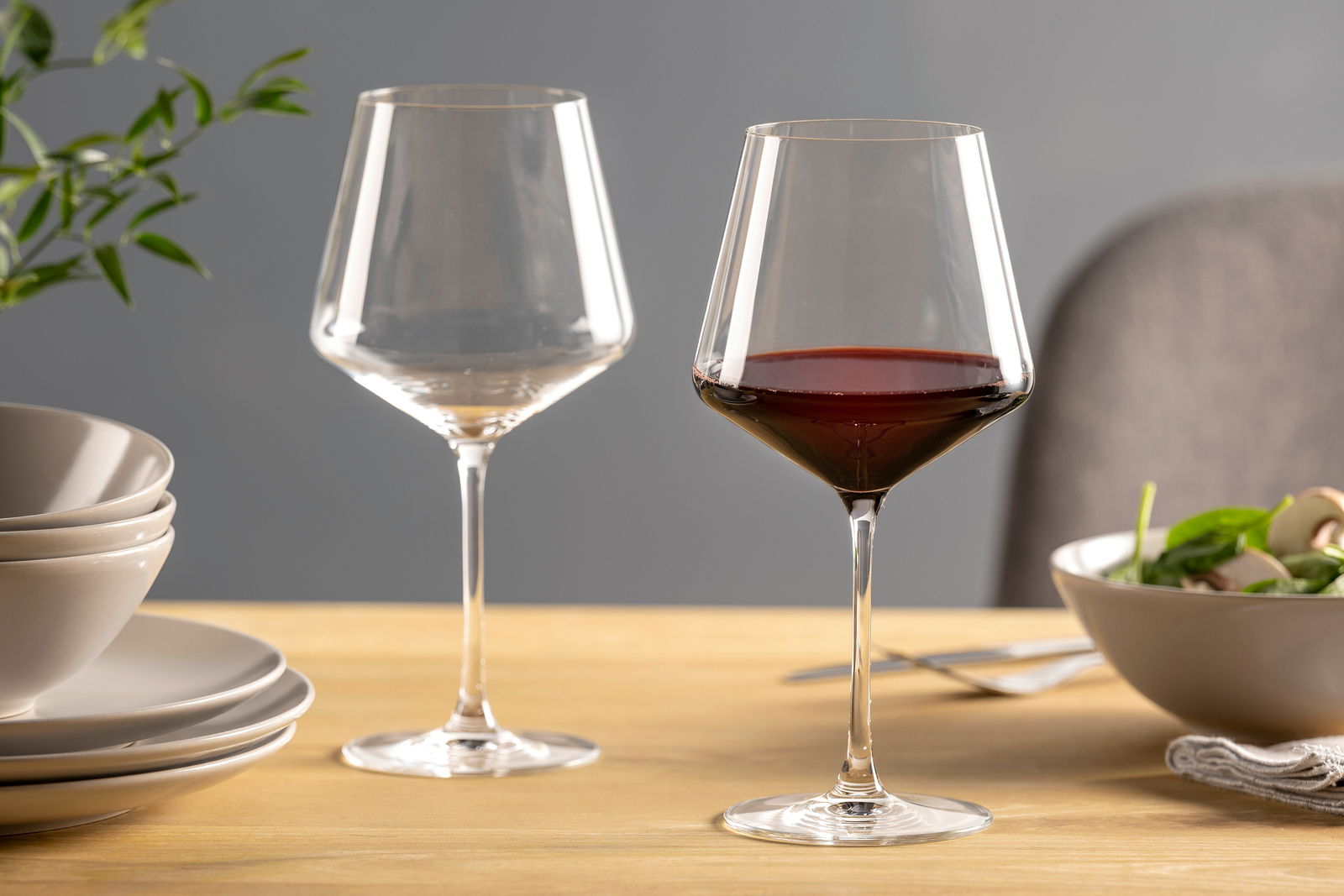 Leonardo Burgunderglas 6er-Set PUCCINI Zwei elegante Burgundergläser auf einem gedeckten Tisch, eines mit Rotwein gefüllt, aus seitlicher Perspektive fotografiert.