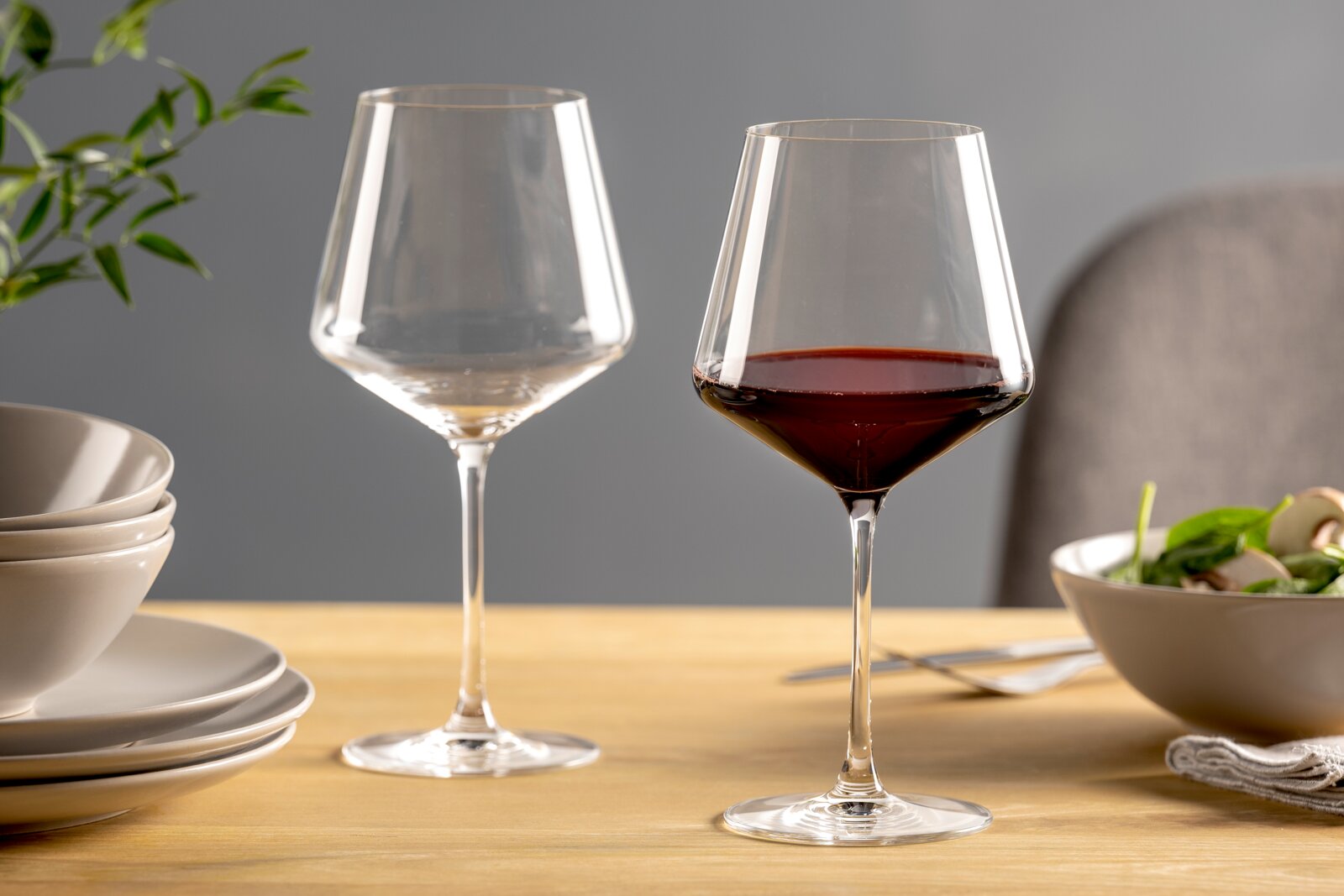 Leonardo Burgunderglas 6er-Set PUCCINI Zwei elegante Burgundergläser auf einem gedeckten Tisch, eines mit Rotwein gefüllt, aus seitlicher Perspektive fotografiert.