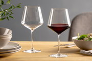 Zwei elegante Burgundergläser auf einem gedeckten Tisch, eines mit Rotwein gefüllt, aus seitlicher Perspektive fotografiert.