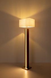 Moderne Stehleuchte mit eckigem Stoffschirm und goldfarbenem Metallfuß, warmes Licht an einer Wand, Frontansicht in leichter Untersicht