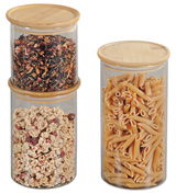 Drei zylindrische Aufbewahrungsgläser mit Bambusdeckeln, gefüllt mit Tee, Müsli und Nudeln, aus der Frontalperspektive.