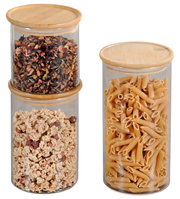 Drei zylindrische Aufbewahrungsgläser mit Bambusdeckeln, gefüllt mit Tee, Müsli und Nudeln, aus der Frontalperspektive.
