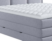 Close-up in schuin vooraanzicht van een wit, gewatteerd topmatras met ritssluiting op een lichtgrijs gestoffeerd boxspringbed
