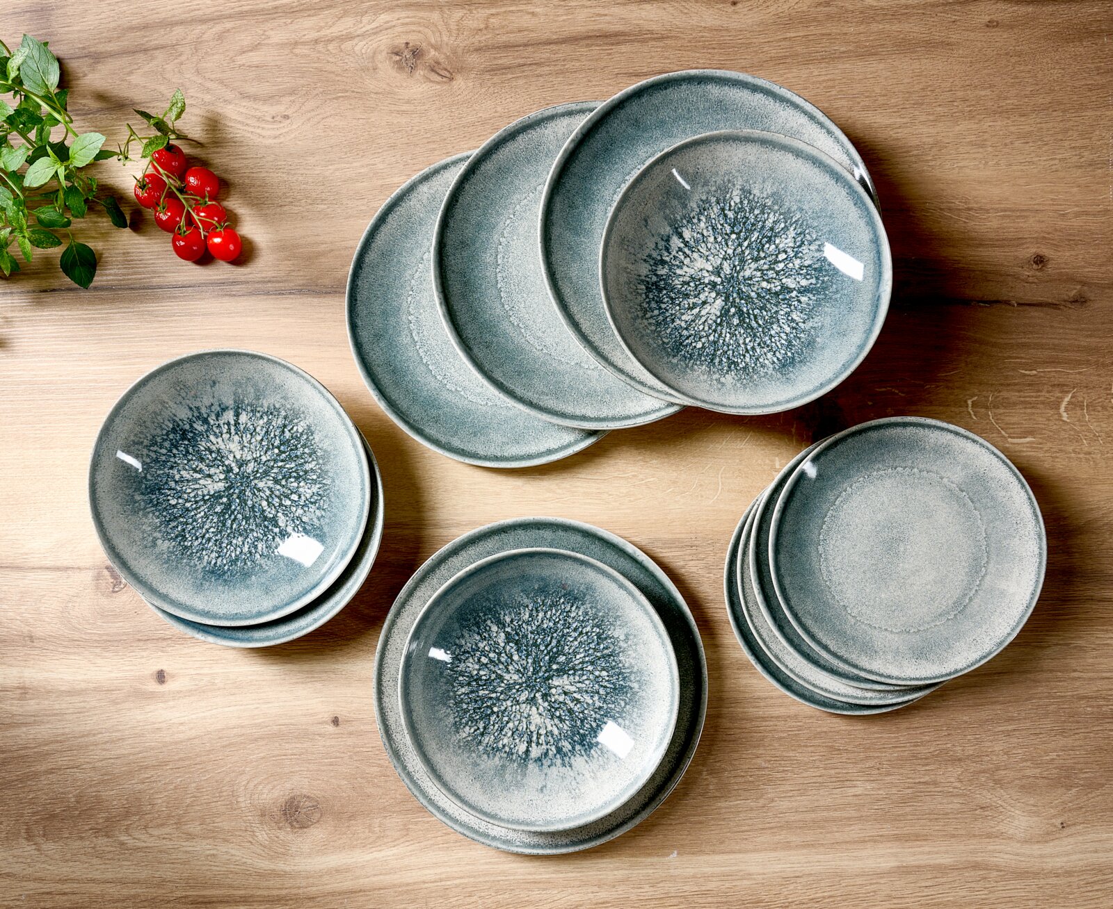 Bovenaanzicht van een stijlvol servies met gestapelde grijsblauwe keramische borden en diepe kommen met een organisch glazuurpatroon op een houten tafel