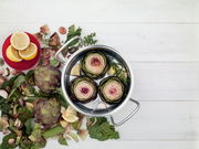 Topf mit Artischocken und Zitronen von oben fotografiert, umgeben von frischen Zutaten auf einem weißen Holztisch.