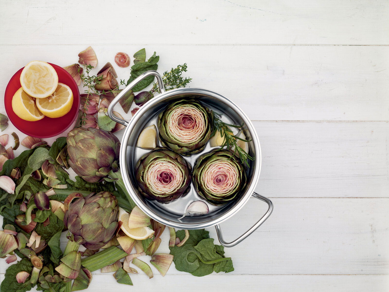 Topf mit Artischocken und Zitronen von oben fotografiert, umgeben von frischen Zutaten auf einem weißen Holztisch.