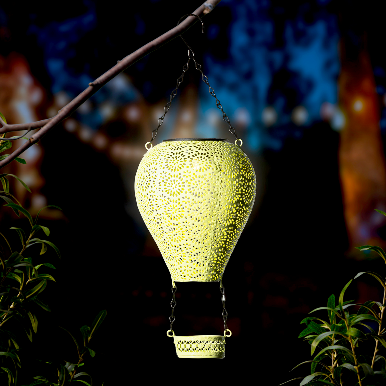 Metallener Solarballon in Form eines Heißluftballons, aufgehängt an einem Ast, leuchtet in der Dämmerung. Perspektive von vorne.