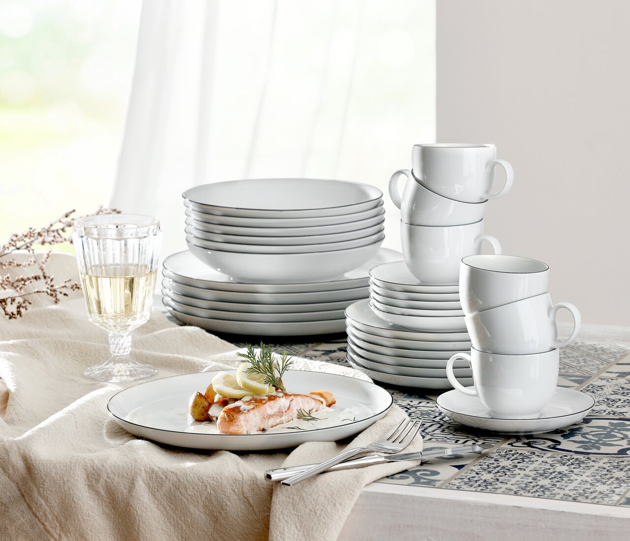 Elegant servies Lido Black, 30-delig, bestaande uit borden, kopjes en schalen, op een gedekte tafel met een glas witte wijn en een bord met een zalmgerecht, zijaanzicht.