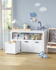 Weißes Kinderzimmer-Aufbewahrungsregal mit zwei Türen und offenem Bücherfach, davor rollbarer Spielzeugkiste; frontale Perspektive in einem hellen Raum mit Deko und Kuscheltieren.