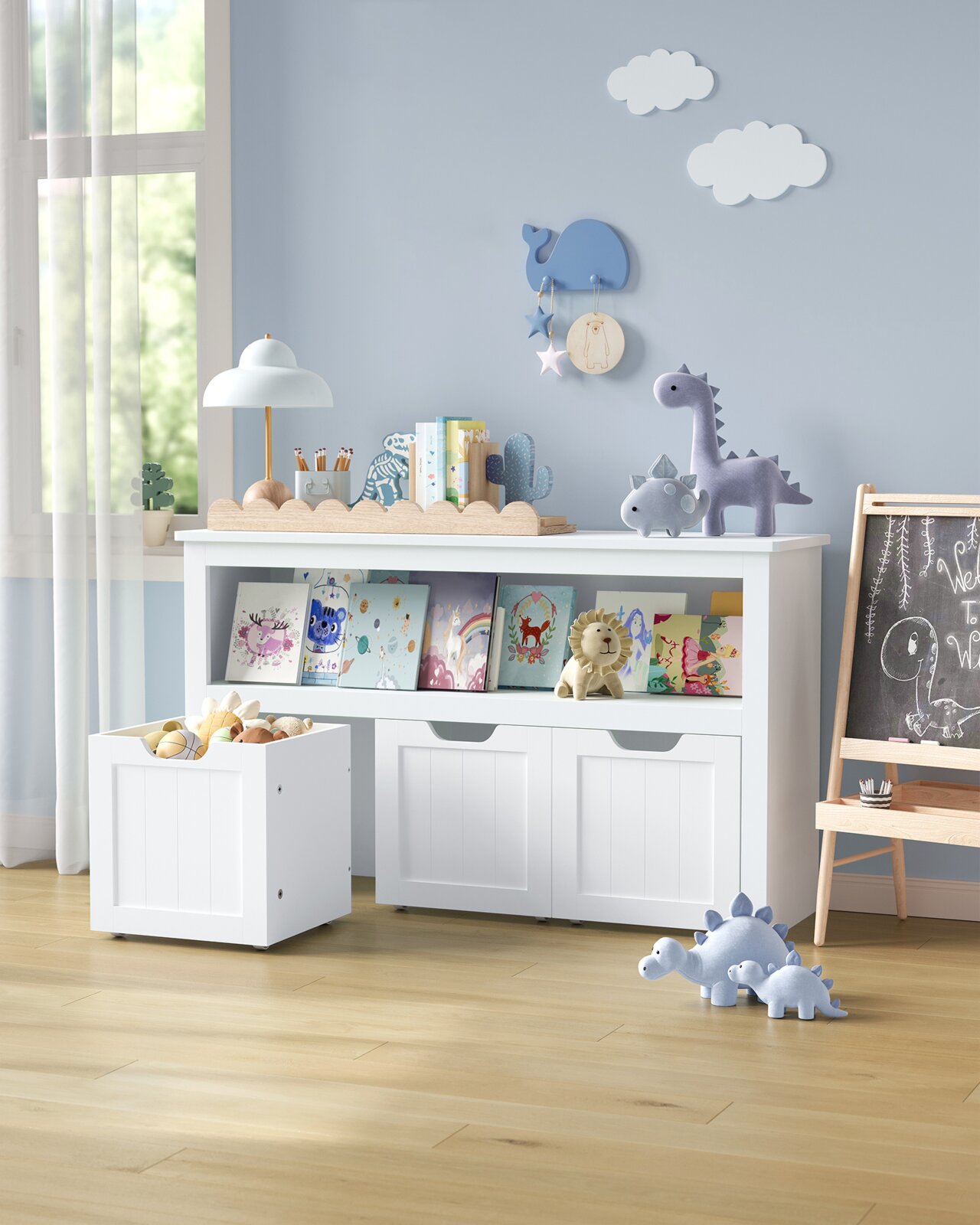 Weißes Kinderzimmer-Aufbewahrungsregal mit zwei Türen und offenem Bücherfach, davor rollbarer Spielzeugkiste; frontale Perspektive in einem hellen Raum mit Deko und Kuscheltieren.