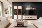 Modernes Wohnzimmer mit weißer Wohnwand in Holzoptik: TV-Lowboard mit Fernseher, zwei Vitrinenschränke, Wandregal und passendem Couchtisch, frontal aus leicht erhöhter Perspektive aufgenommen.