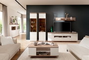 Modernes Wohnzimmer mit weißer Wohnwand in Holzoptik: TV-Lowboard mit Fernseher, zwei Vitrinenschränke, Wandregal und passendem Couchtisch, frontal aus leicht erhöhter Perspektive aufgenommen.