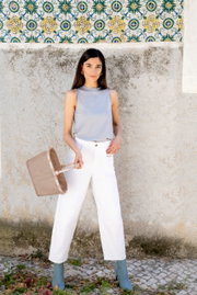 Een vrouw in een witte broek en een grijze top houdt een klein, beige mandje vast, gezien vanuit een zijaanzicht.