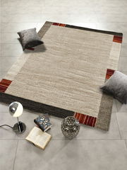 Moderner Teppich in Beige- und Brauntönen mit roten Akzenten, dekoriert mit Kissen, Büchern und einer Lampe, aus der Vogelperspektive fotografiert.