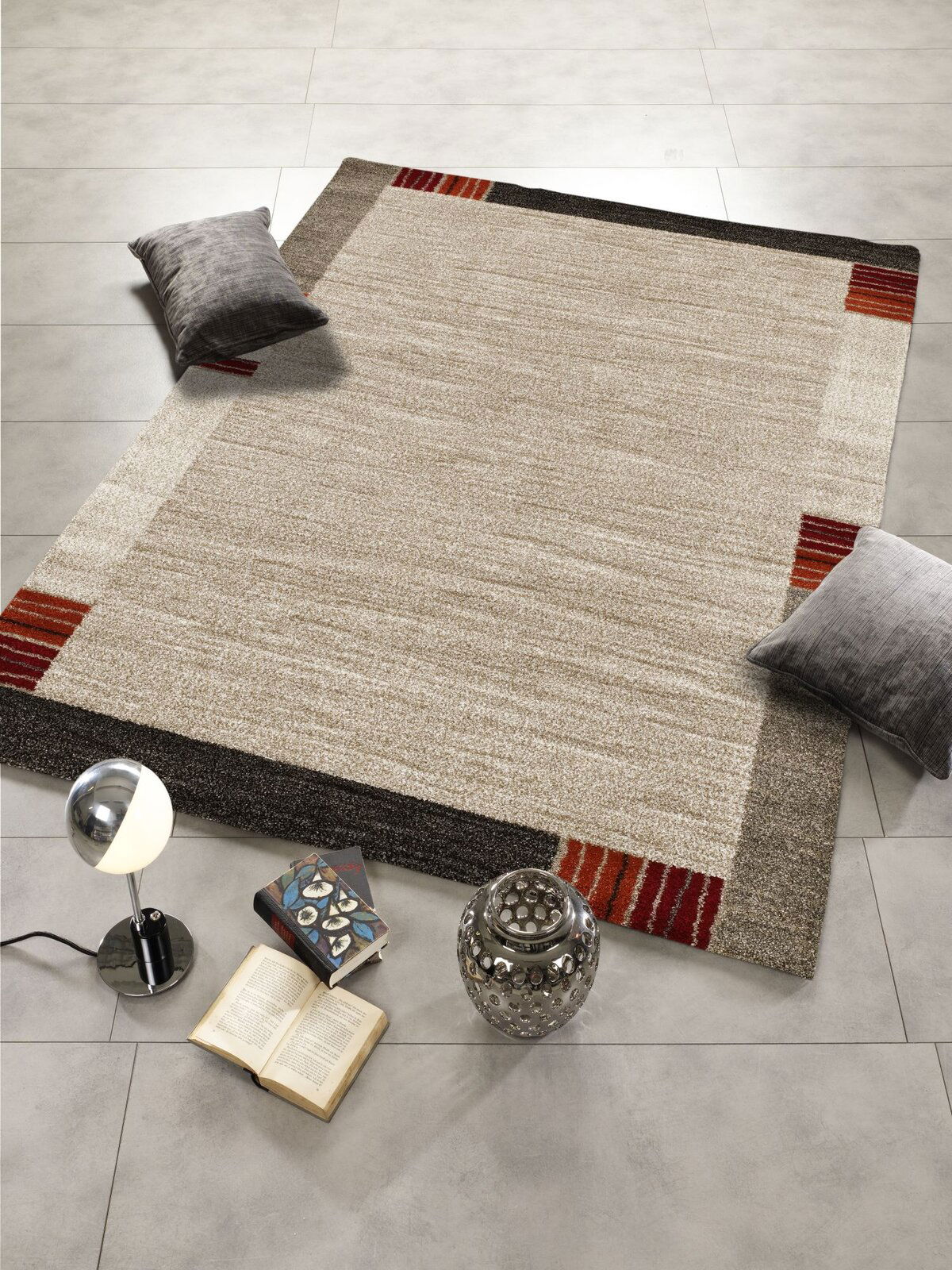 Moderner Teppich in Beige- und Brauntönen mit roten Akzenten, dekoriert mit Kissen, Büchern und einer Lampe, aus der Vogelperspektive fotografiert.
