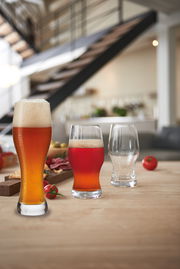 Drei Biergläser auf einem Tisch in einem modernen Raum, gefüllt mit verschiedenen Biersorten, im Vordergrund ein Weizenbierglas mit Schaumkrone.