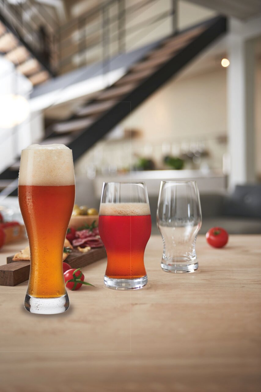 Drei Biergläser auf einem Tisch in einem modernen Raum, gefüllt mit verschiedenen Biersorten, im Vordergrund ein Weizenbierglas mit Schaumkrone.