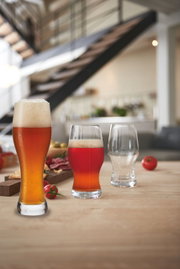 Drei Biergläser auf einem Tisch in einem modernen Raum, gefüllt mit verschiedenen Biersorten, im Vordergrund ein Weizenbierglas mit Schaumkrone.