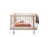 Beiges Babybett mit Gitterseiten und Matratze, mit rosa Decke und Kuscheltier, Frontansicht auf weißem Hintergrund