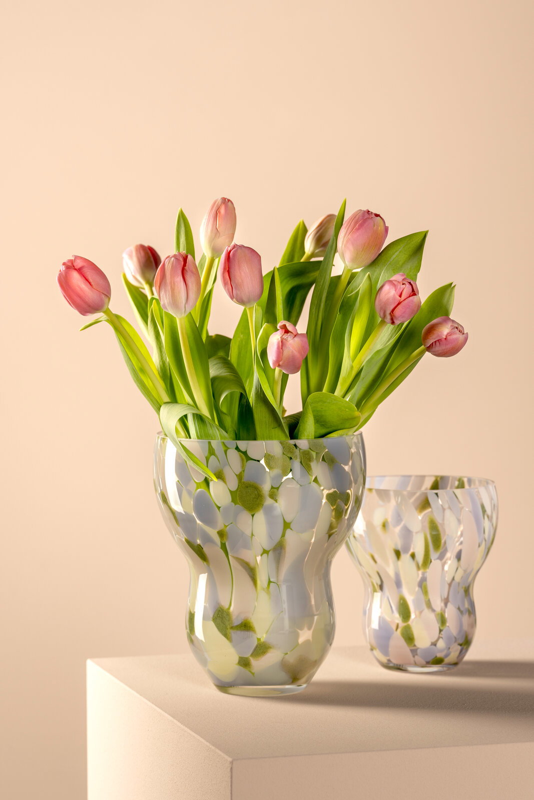 Zwei Vasen mit buntem, organischem Muster, gefüllt mit rosa Tulpen, auf einem Podest aus seitlicher Perspektive.