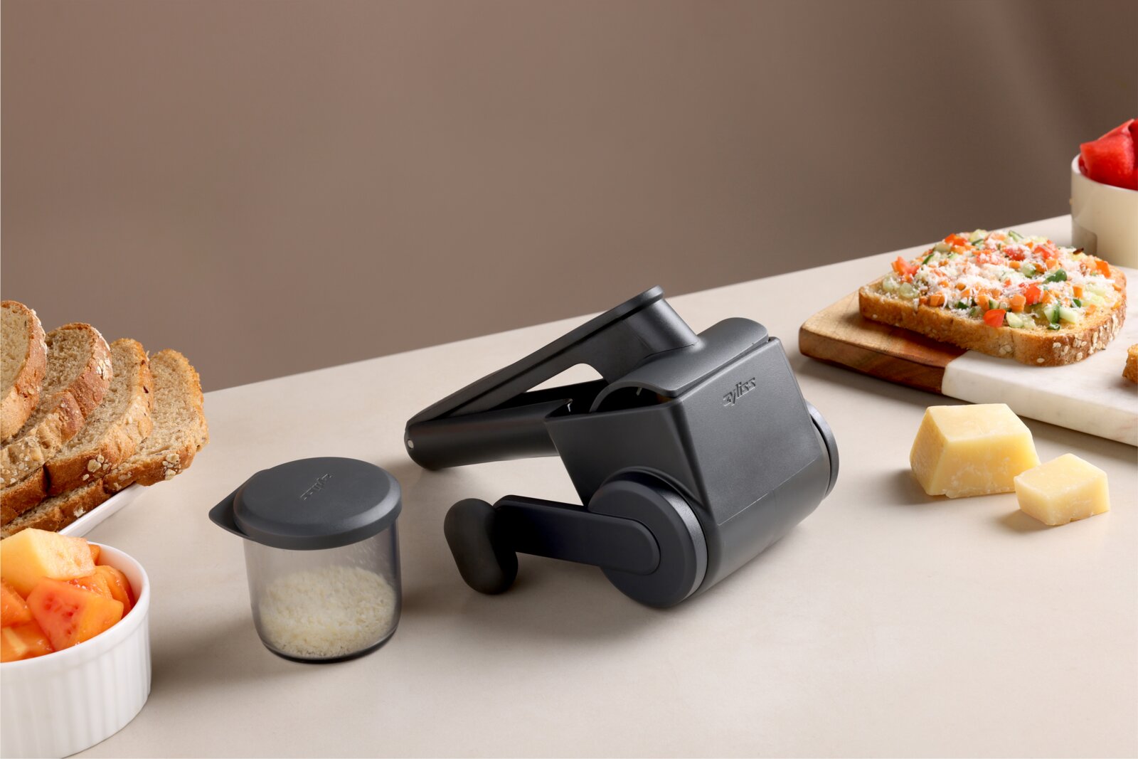Schwarze manuelle Käsereibe mit Kurbel und Auffangbehälter auf Küchenarbeitsplatte, seitliche Perspektive (3/4-Ansicht) mit Brot, belegtem Toast und Käsewürfeln im Hintergrund