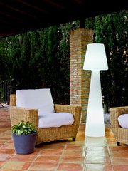 Leuchtende weiße Outdoor-Stehleuchte in modernem Design auf einer überdachten Terrasse neben Rattan-Loungesessel mit weißen Polstern, Frontansicht auf Augenhöhe
