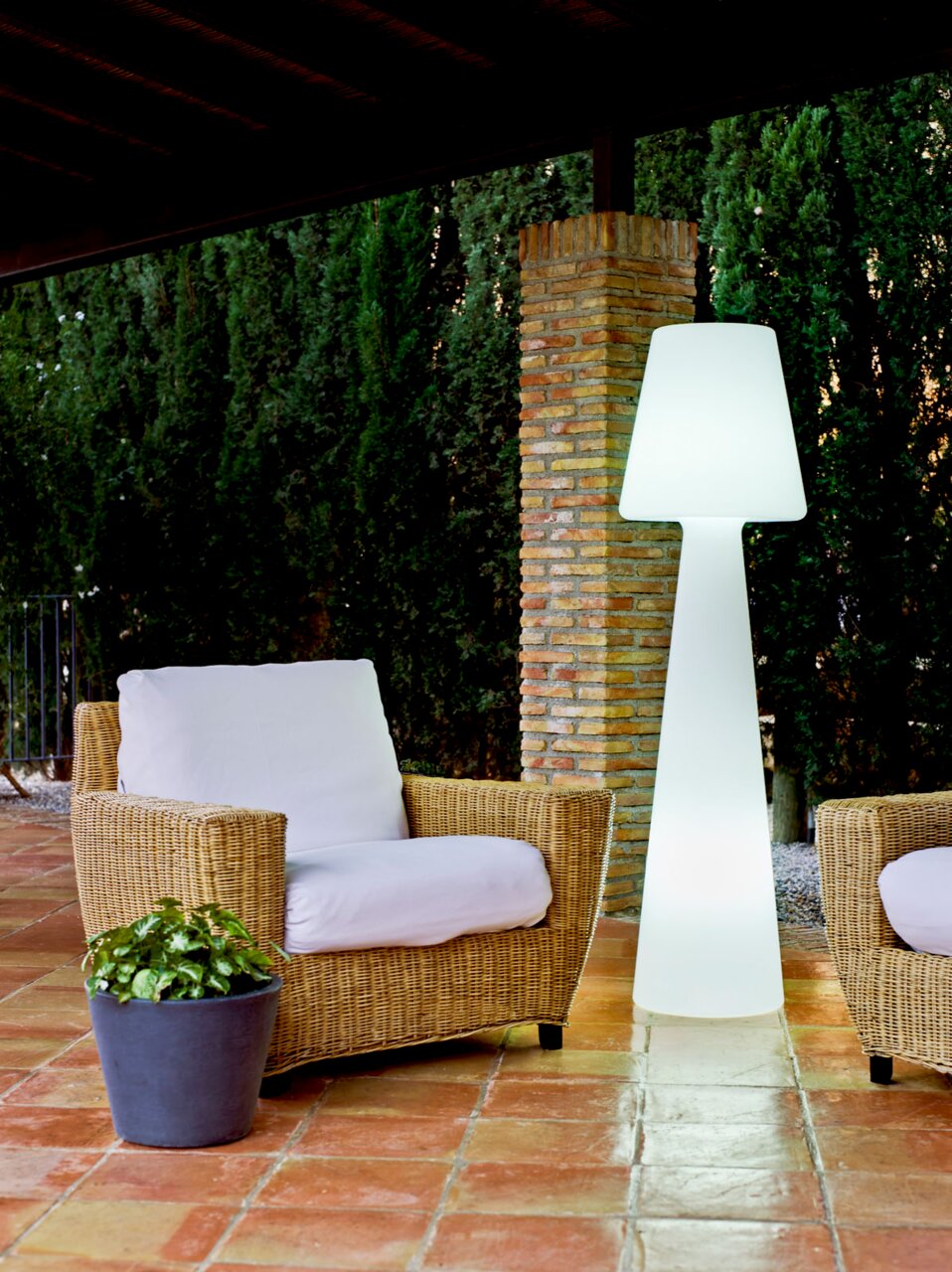 Leuchtende weiße Outdoor-Stehleuchte in modernem Design auf einer überdachten Terrasse neben Rattan-Loungesessel mit weißen Polstern, Frontansicht auf Augenhöhe