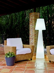 Leuchtende weiße Outdoor-Stehleuchte in modernem Design auf einer überdachten Terrasse neben Rattan-Loungesessel mit weißen Polstern, Frontansicht auf Augenhöhe