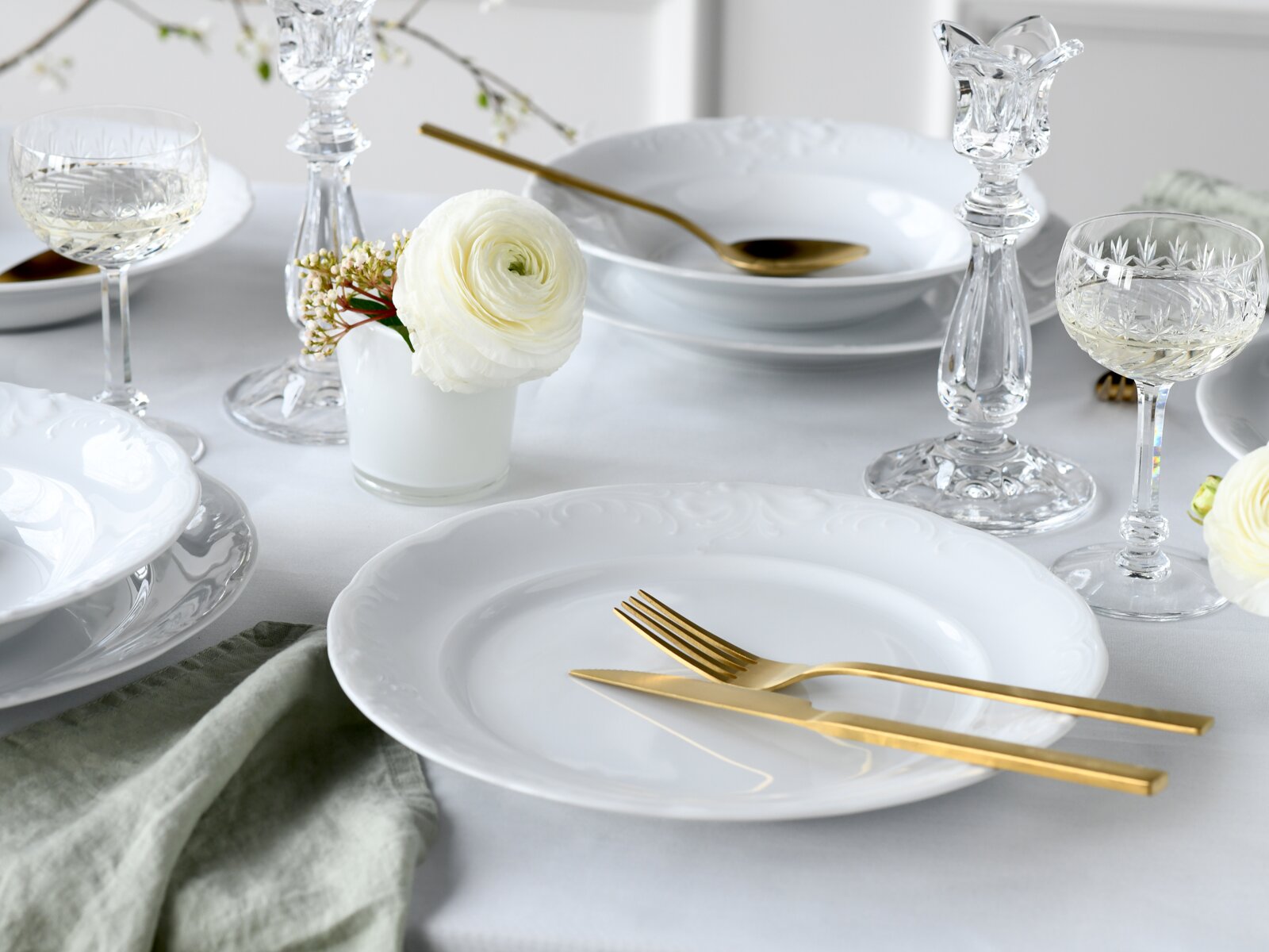 Elegant gedeckter Tisch mit weißen Porzellantellern, goldfarbenem Besteck und Kristallgläsern, dekoriert mit weißen Blumen – Perspektive: leicht von oben, schräger Blick auf das Gedeck.