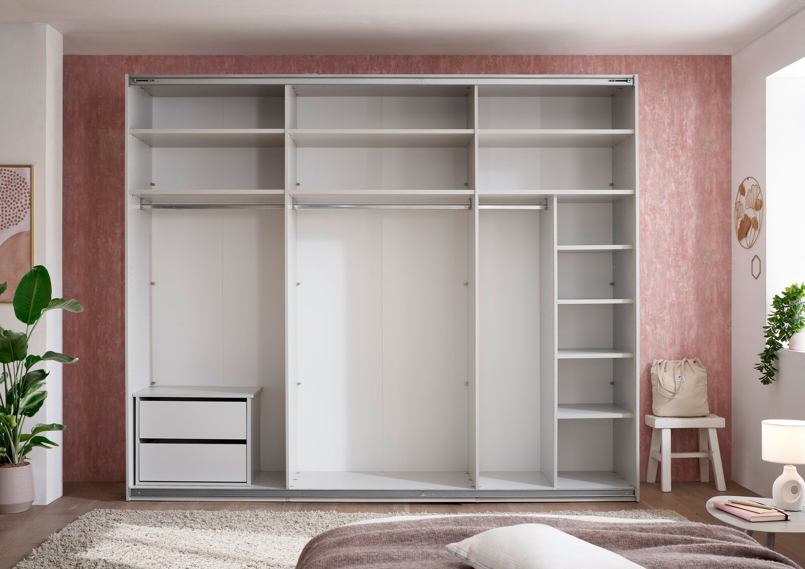 Frontansicht eines offenen, weißen Einbauschranks/Schranksystems mit mehreren Fächern, zwei Kleiderstangen und einem Schubladenelement links in einem Schlafzimmer.