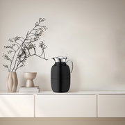 Schwarze Isolierkanne Juwel auf einem weißen Regal, seitliche Perspektive, neben dekorativen Vasen und Zweigen.