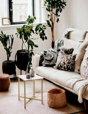 Helles Wohnzimmer mit weißem Sofa, gemusterten Kissen und Plaid, goldfarbenem Beistelltisch mit weißer Marmoroptik-Platte sowie großen Zimmerpflanzen am Fenster, fotografiert aus leicht erhöhter schräger Frontperspektive.