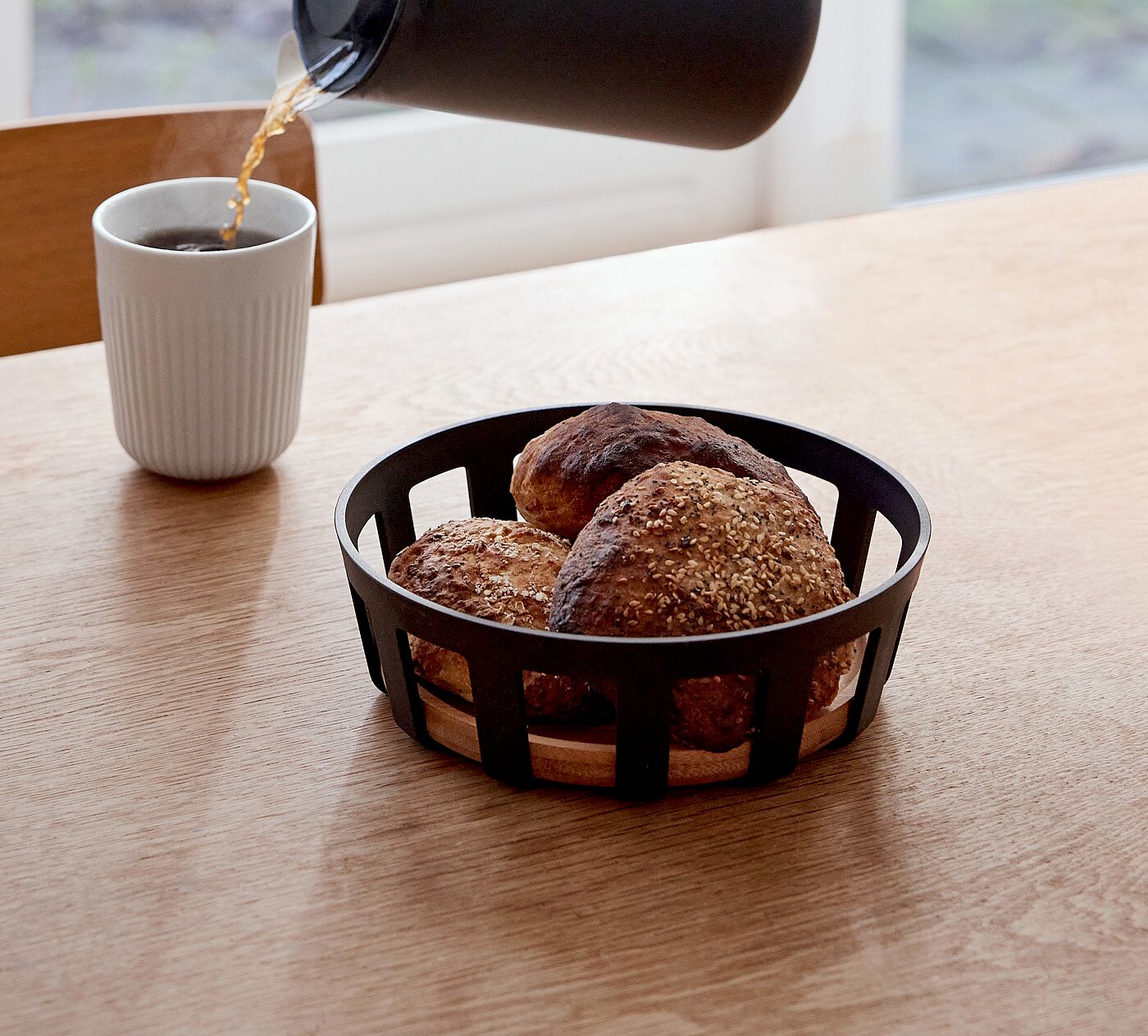 Zwart metalen broodmandje met houten bodem en drie broodjes op een houten tafel, gefotografeerd schuin van voren; op de achtergrond wordt koffie in een witte mok geschonken.
