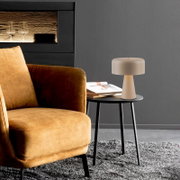 Beige Tischlampe mit breitem zylindrischem Schirm auf rundem schwarzem Beistelltisch neben cognacfarbenem Sessel in modernem Wohnzimmer, Frontansicht in leicht schräger Perspektive von links