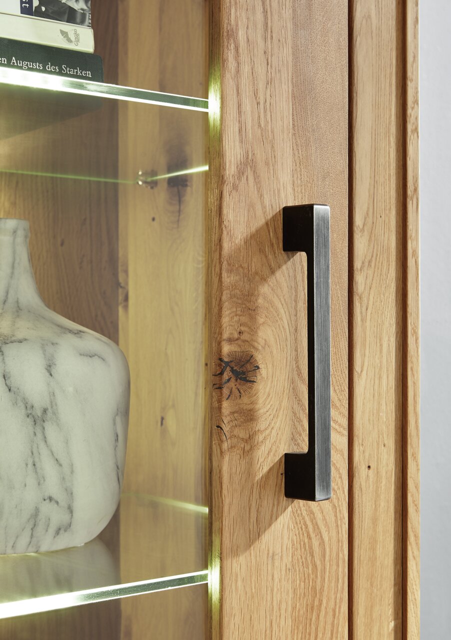 Nahaufnahme einer Vitrine aus Holz mit Glastür, Metallgriff und beleuchteten Glasregalen, Perspektive von der Seite.