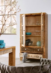 Holzvitrine mit Glastüren, dekoriert mit Geschirr und Gläsern, in einem modernen Wohnzimmer mit Blick auf ein großes Fenster.