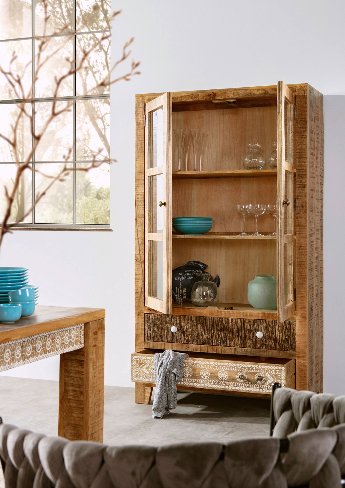 Holzvitrine mit Glastüren, dekoriert mit Geschirr und Gläsern, in einem modernen Wohnzimmer mit Blick auf ein großes Fenster.