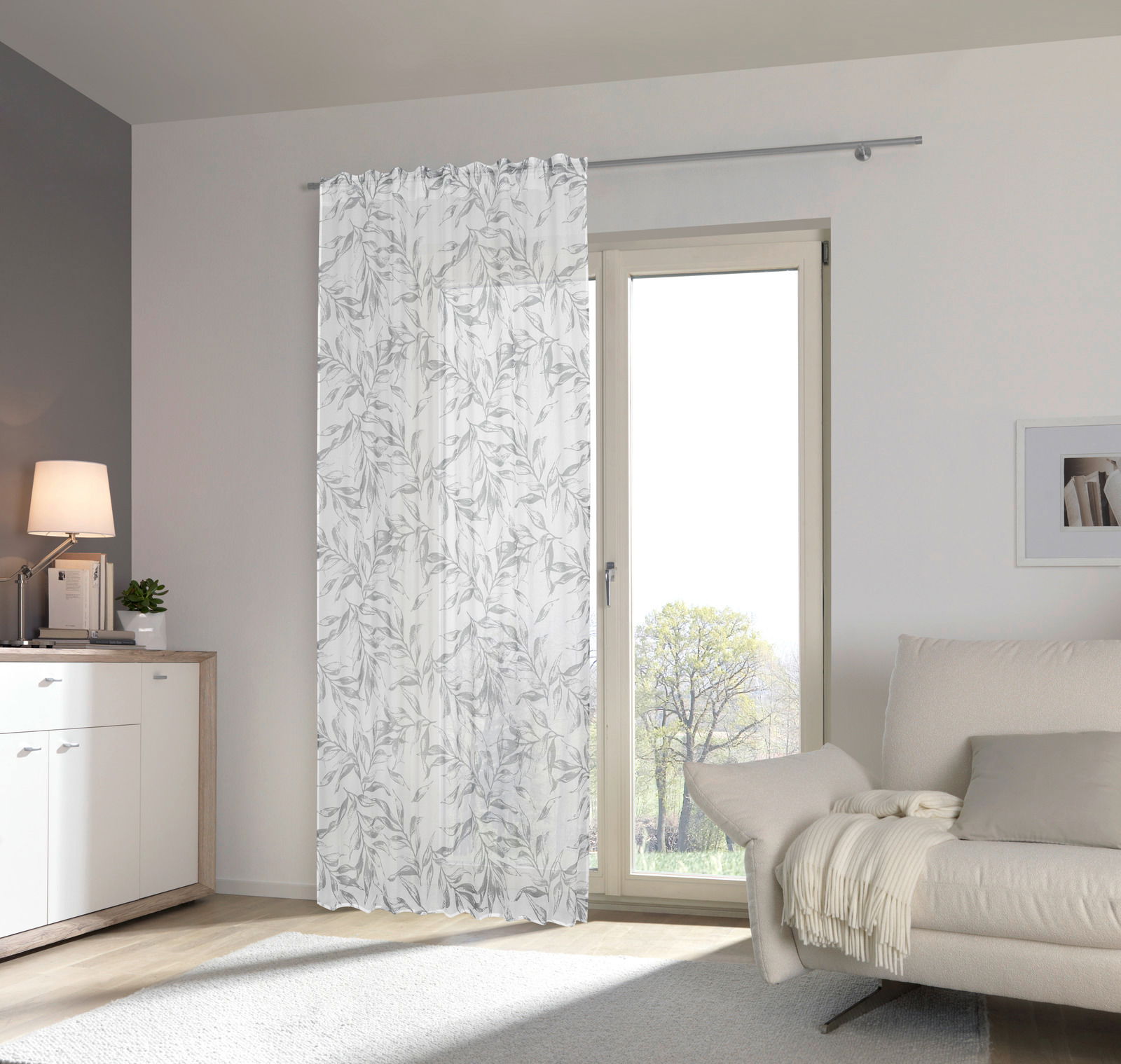 Fertigvorhang Flora 140x245 in einem modernen Wohnzimmer, halbtransparent mit floralem Muster, aufgehängt an einer Gardinenstange vor einer großen Glastür, seitliche Perspektive.